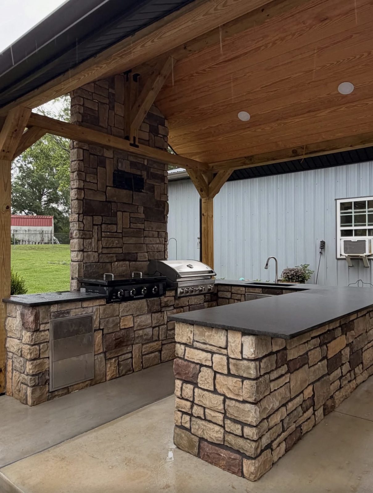 Covered outdoor kitchen and bar with a full stone base and dark slab countertops.