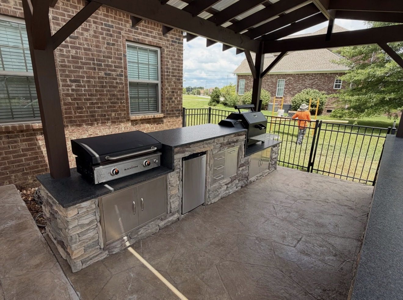 Outdoor kitchen with a stacked stone base and dark countertops under a pergola on a stamped concrete patio.